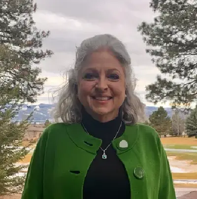 A woman in green jacket standing outside near trees.