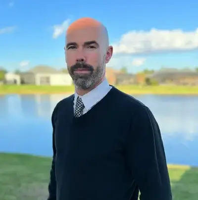 A man with bald head and beard standing in front of water.