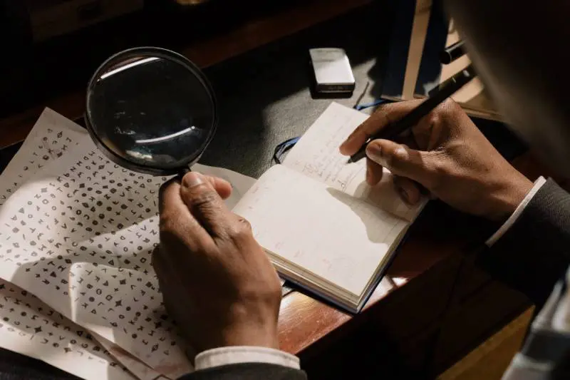 A person writing in a notebook with a magnifying glass.