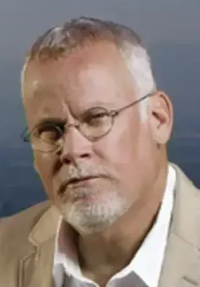 A man with white hair and glasses in front of the ocean.